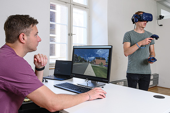 En primer plano a la izquierda, un hombre sentado en un escritorio con una computadora observa a un hombre a la derecha con un visor de realidad virtual.