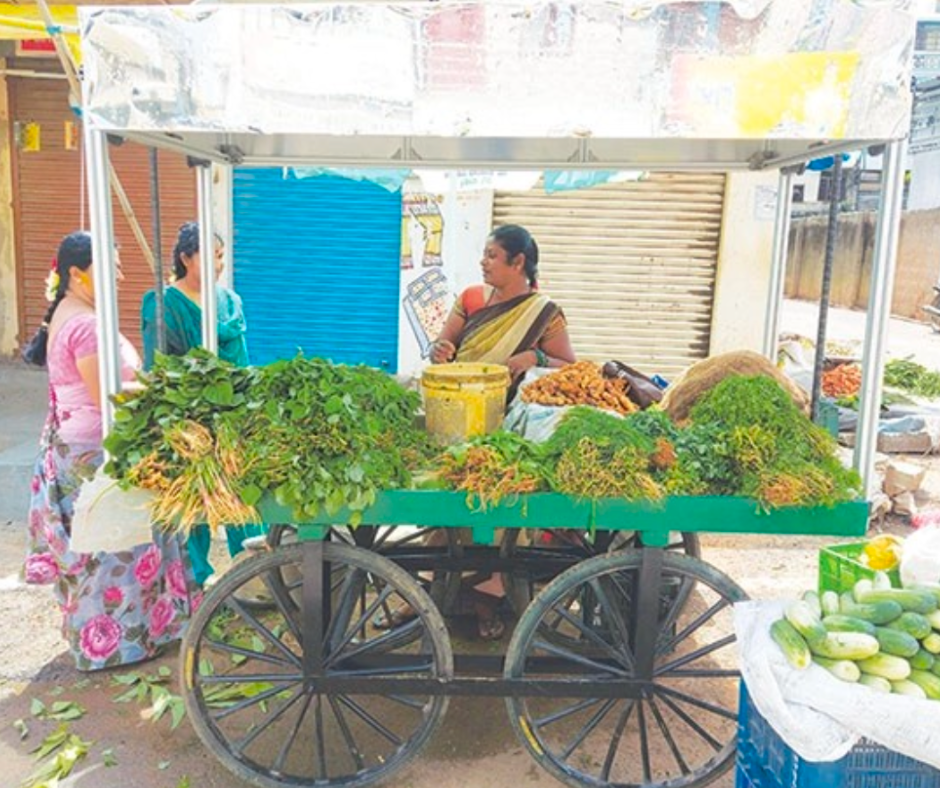 Imagen de  un carrito de verduras en la calle. Está lleno de hojas verdes, pepinos y otros vegetales frescos. Una mujer vendedora, con ropa tradicional, habla con dos clientas que observan los productos. Al fondo hay locales cerrados y la escena ocurre a plena luz del día.